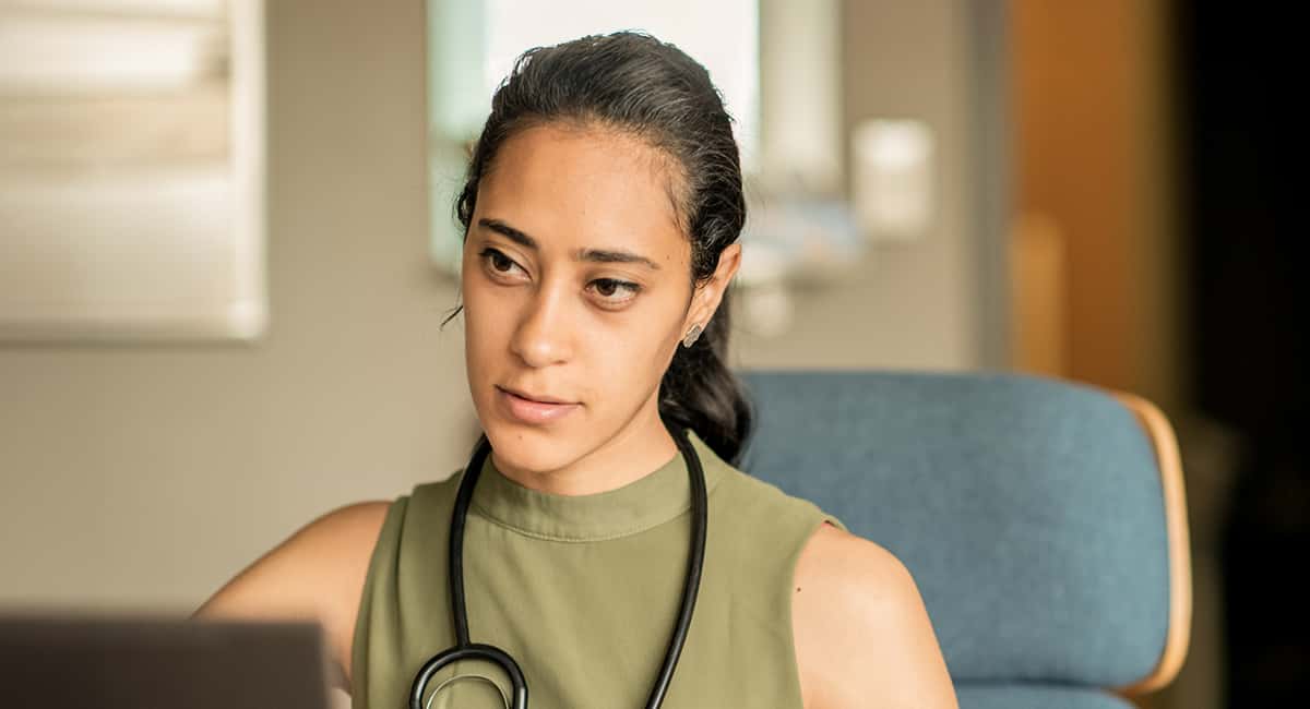 A professional-looking, dark-haired doctor with a stethoscope around her neck, wearing a green blouse, looks intently at a laptop screen. The background is a slightly blurred office setting.