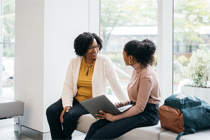 Two women are sitting on a bench in a bright, modern waiting area or lounge. An older woman with glasses is smiling at a younger woman who is holding a laptop.