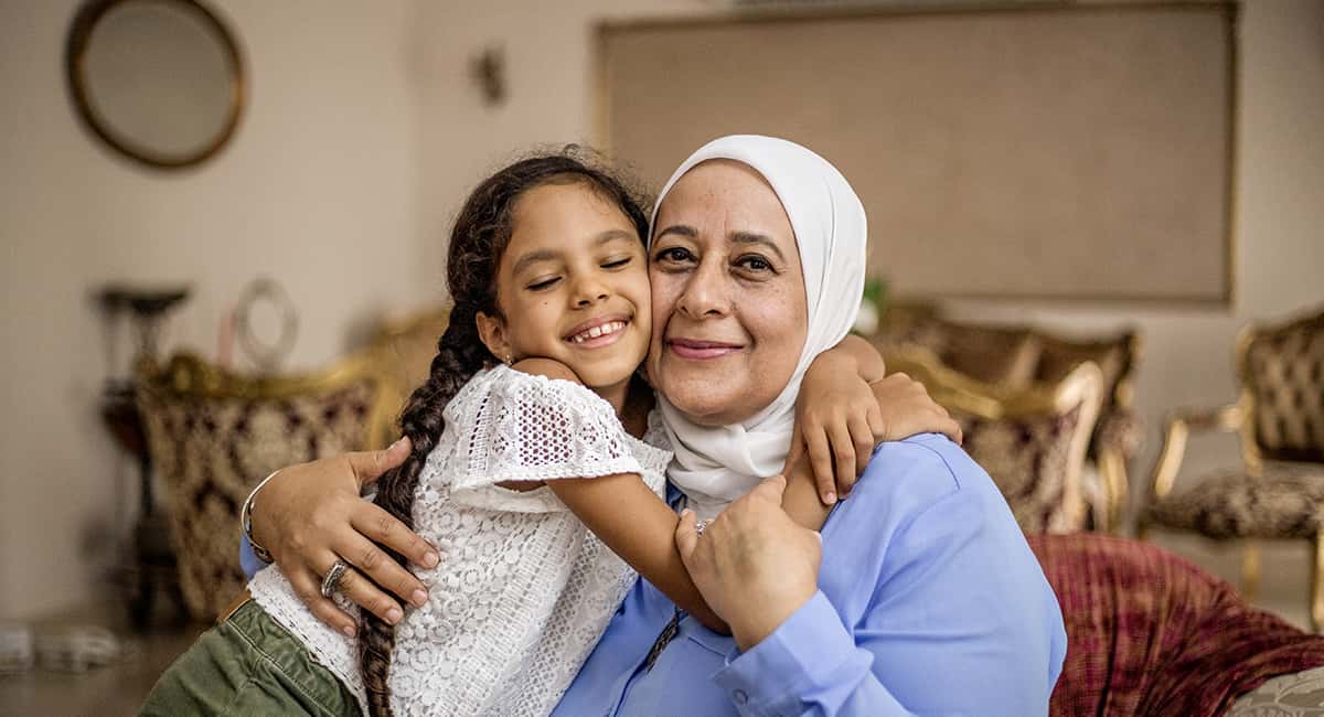 A smiling elder woman is being hugged by a child in a home environment.