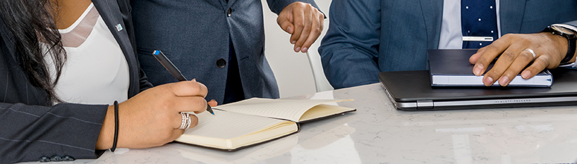 Three people's hands are shown at a desk with notebooks out and pen poised.