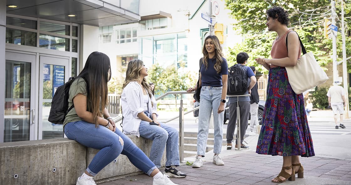 Four students are hanging out outside the George Vari Engineering and Computing Centre. Two students are sitting and two are standing. They are looking at one student who is smiling and talking. 
