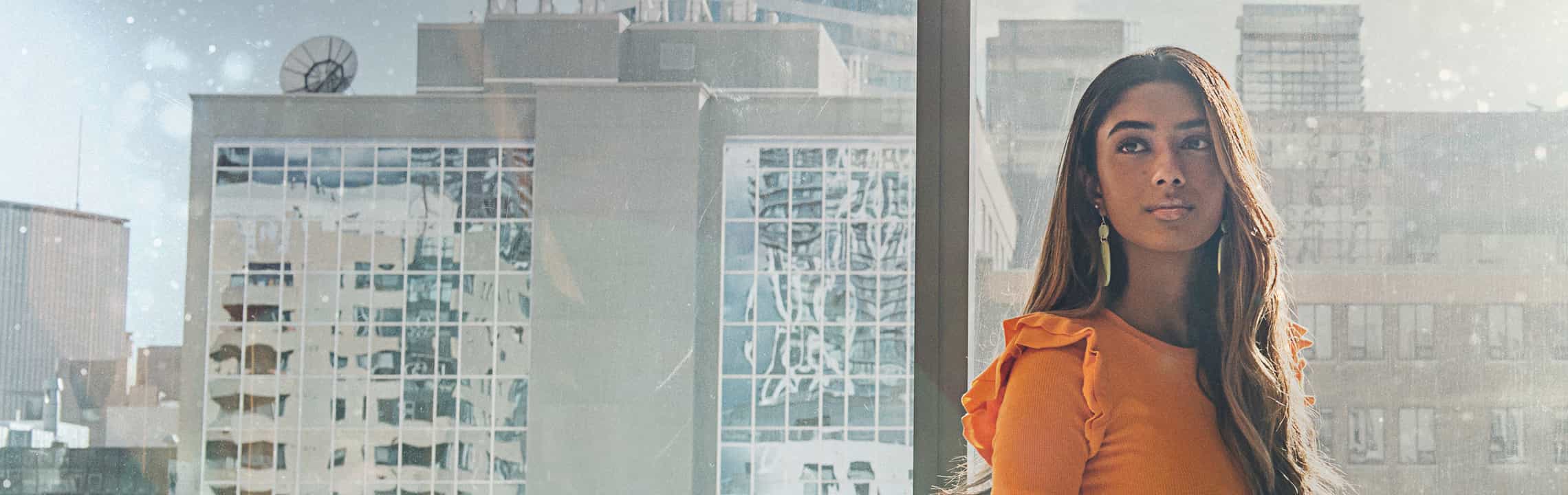 A close up of a female student, standing in front of a glass window with a view of Toronto buildings. She is looking away from the camera.