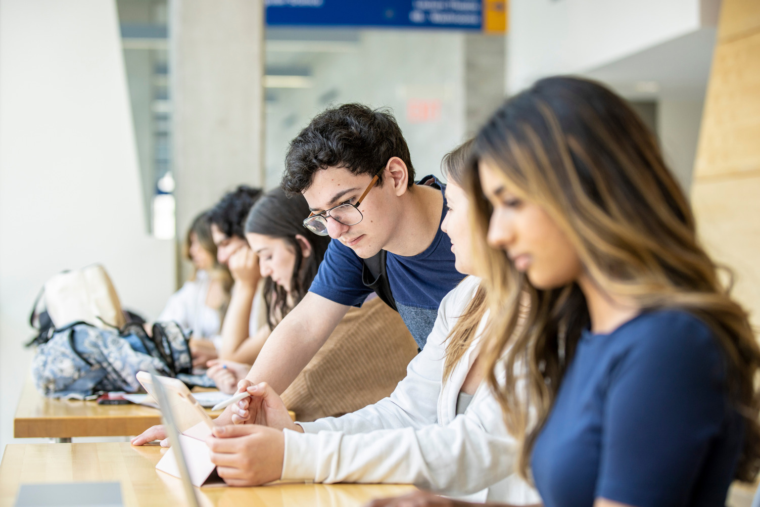 A male student leaning to assist a female student with her studies. Other students can be seen in the background.   