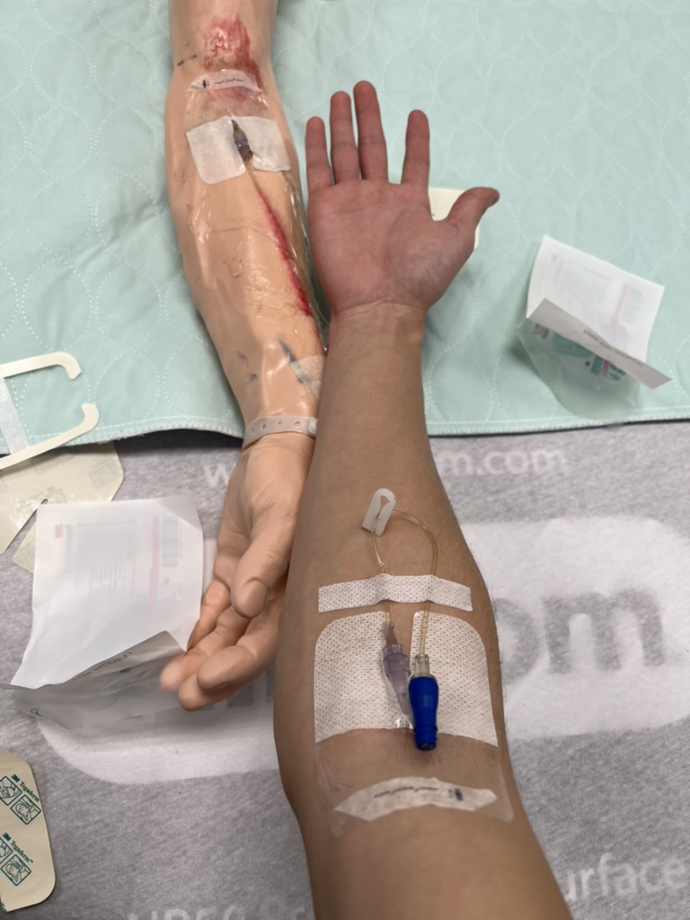 Nick's arm extended by a medical mannequin arm, both have IV drips inserted 