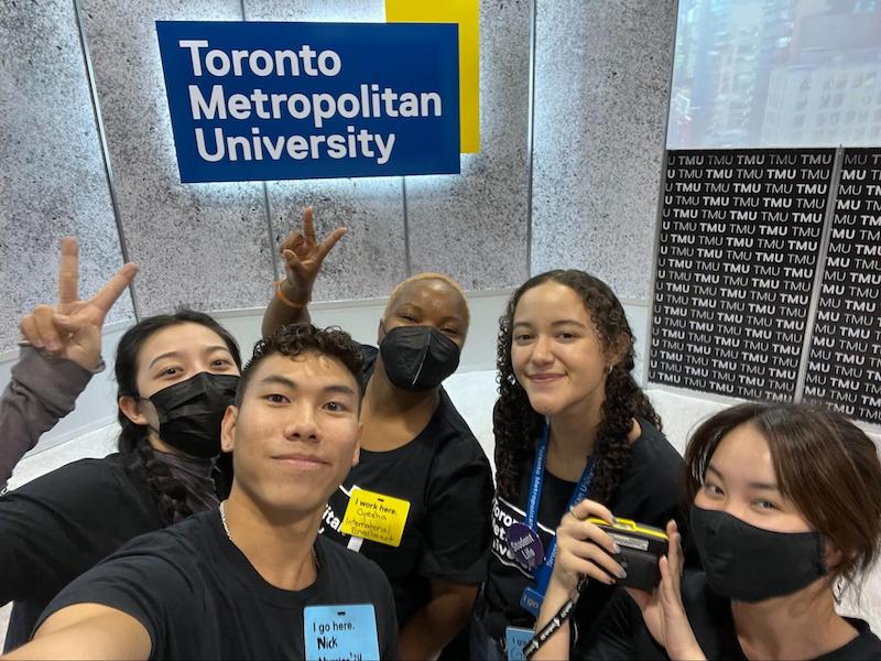 Giuli and her coworkers pose for a selfie. Two team members are making a peace sign in the air. A plaque of the TMU logo is in the back ground