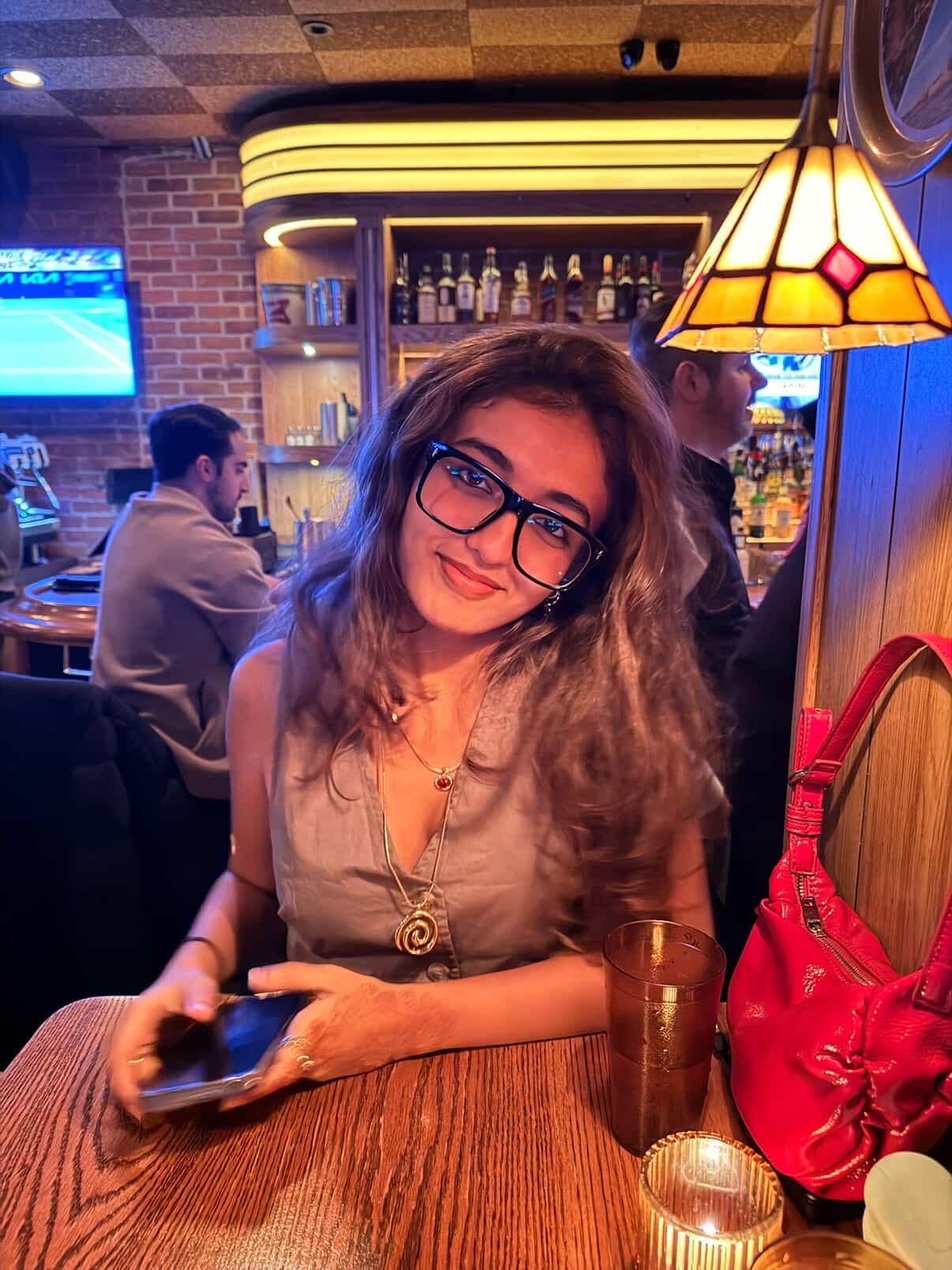 A young woman with glasses and wavy brown hair smiling at a bar table