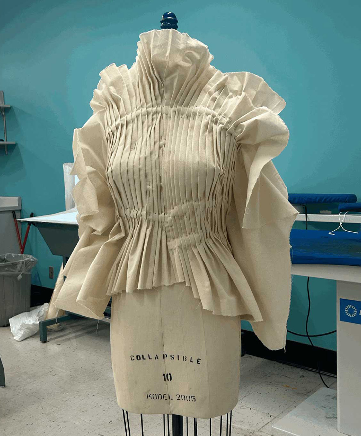 A pleated cream fabric garment with ruffled sleeves on a dress form in a fashion studio