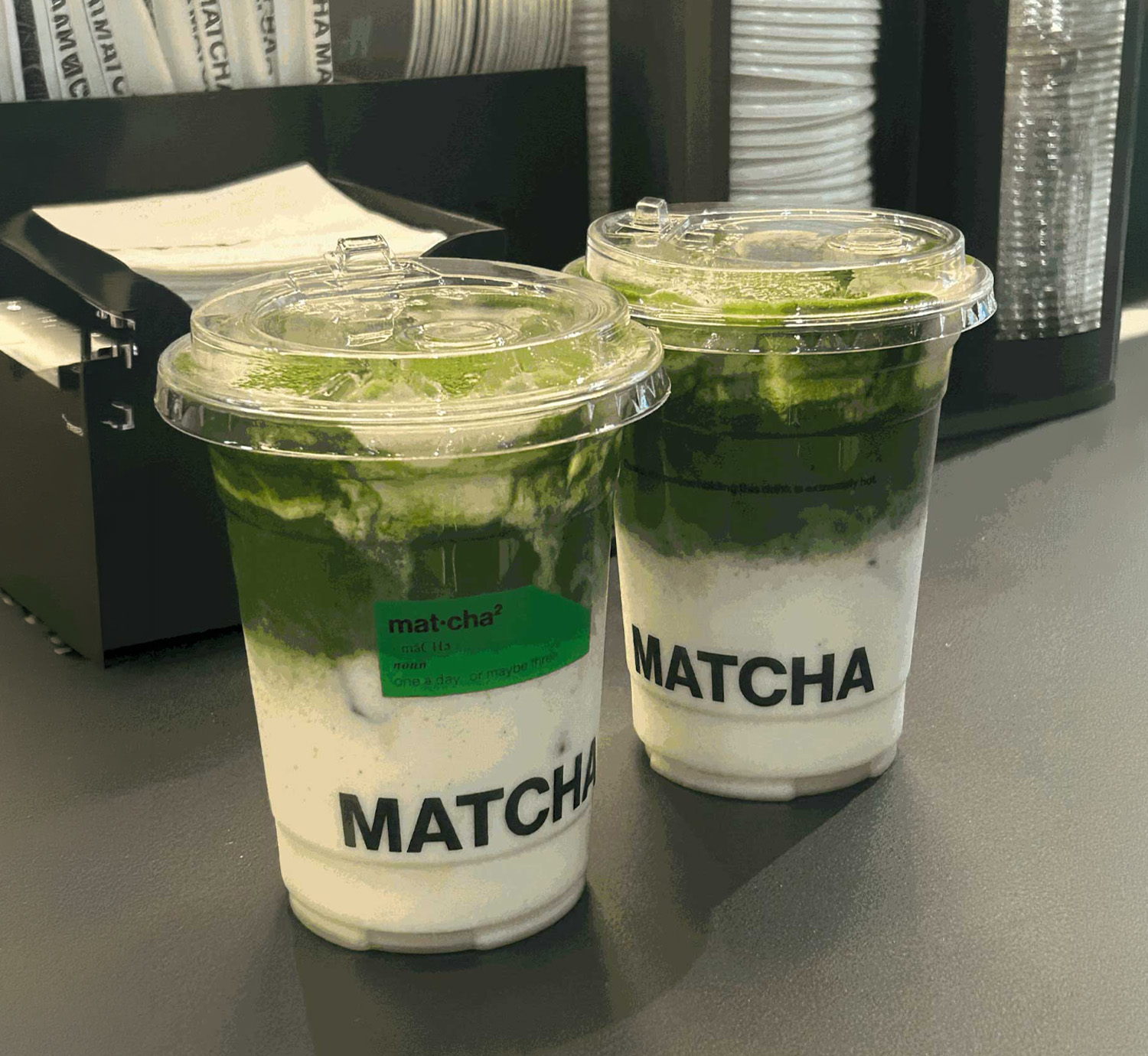 Two iced matcha lattes in branded clear plastic cups on a black table