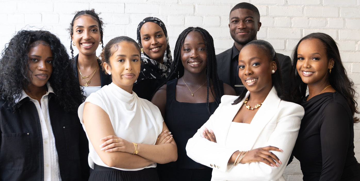 A group of eight young professionals posing together in front of a white brick wall, dressed in smart casual and business attire.