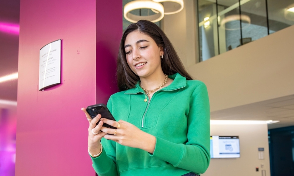 A student is standing at the Service Hub, looking at her cell phone and smiling. 