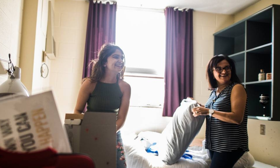 A parent helps her daughter settle in to her new room on campus. They are smiling and talking to a third person not pictured.