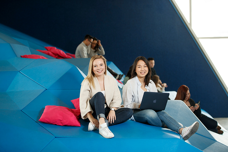 Two students are studying with a laptop in Sandbox by DMZ in SLC. They are looking at the camera. Other students can be seen in the background.