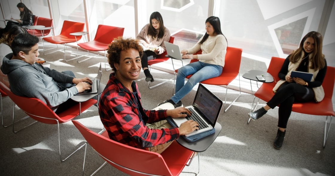 Five students are sitting in the Student Learning Centre (SLC). They are working on their laptops. One student is looking at the camera, smiling. 