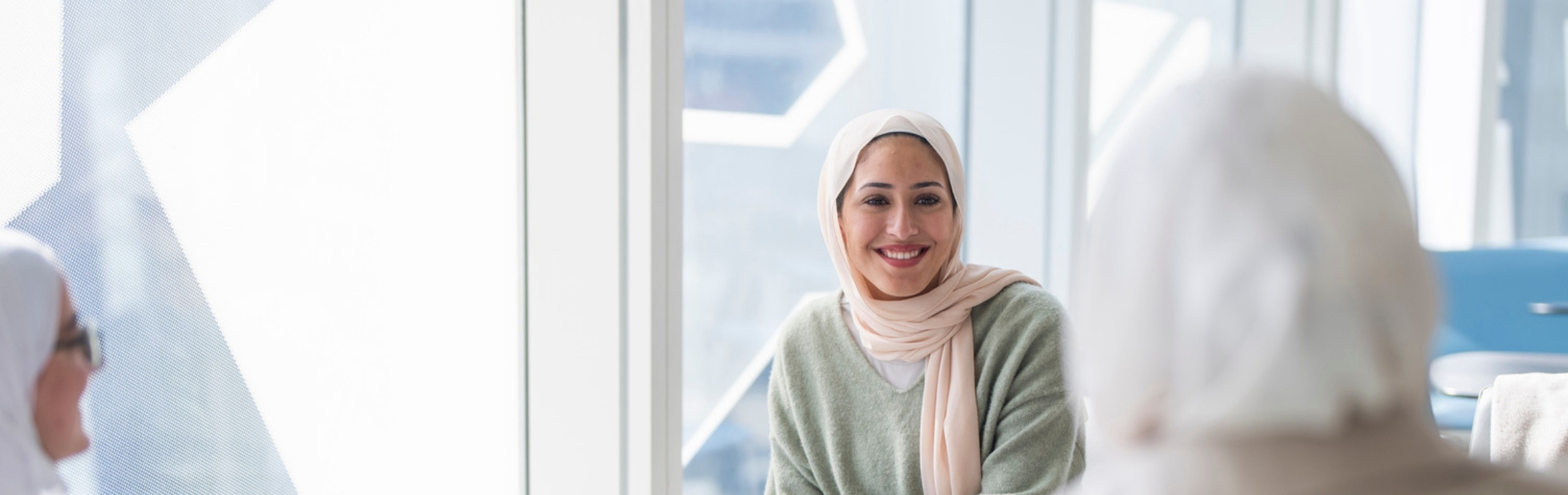 Smiling woman wearing a green sweater sitting in a bright modern room, engaging in conversation with others near large windows.