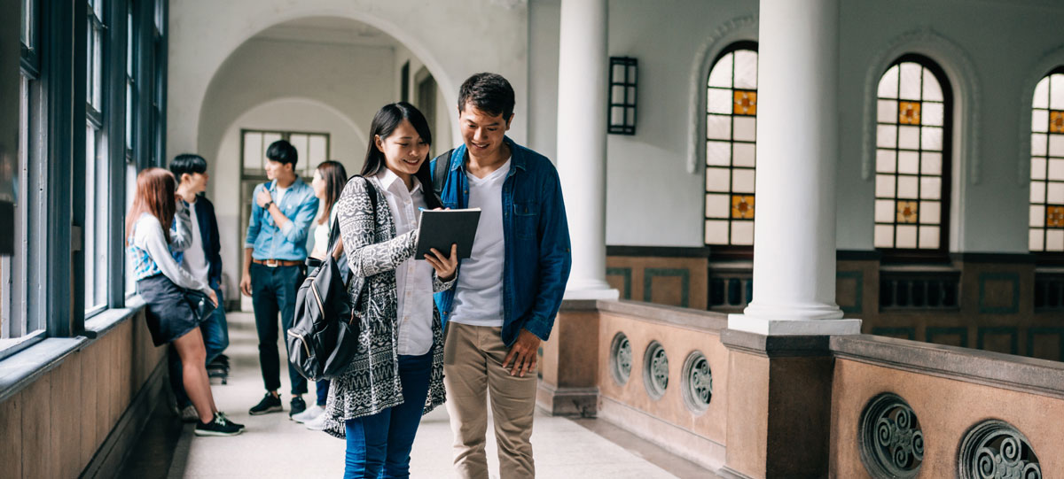 Students standing and talking in a bright hallway with arched windows and white columns, as two people in the foreground look at a tablet together and smile.