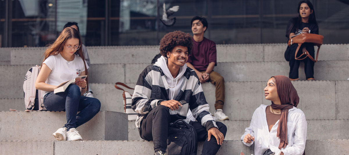 Group of university students sitting and chatting on outdoor concrete steps, with a sunny campus setting.