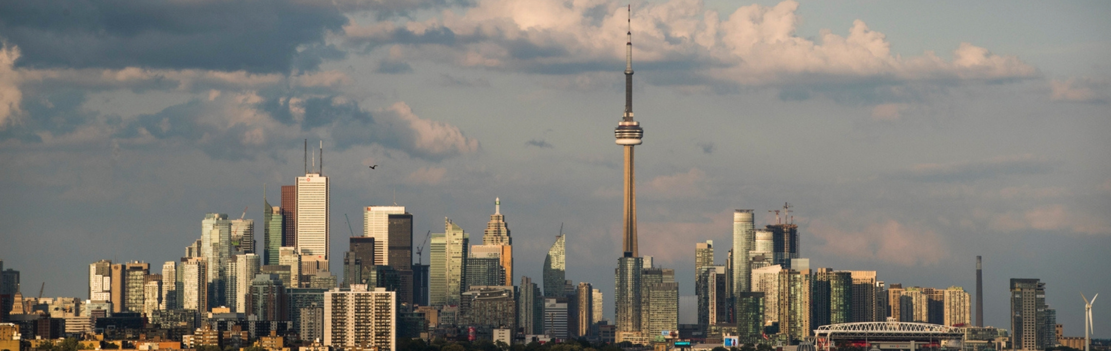 Wide-angle view of downtown Toronto’s skyline under a partly cloudy sky, featuring the CN Tower prominently surrounded by high-rise office and residential buildings.