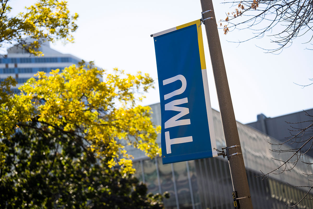 A TMU banner hangs on a light pole, with trees and campus buildings in the background