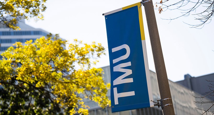 A TMU banner hangs on a pole on campus on a sunny day.