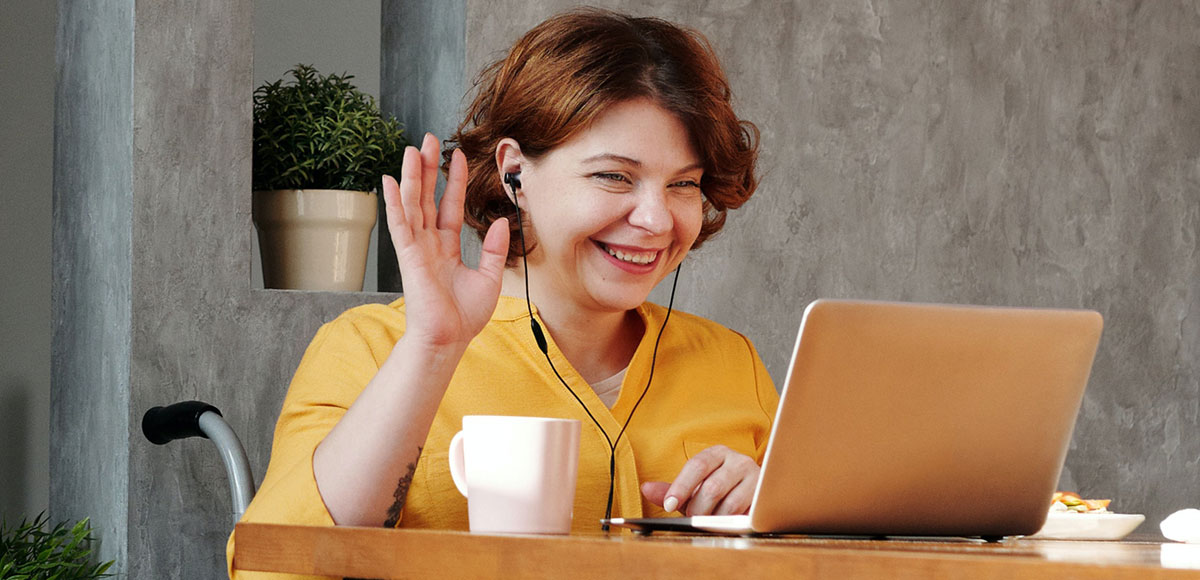 A woman sitting at a table and smiling and waving at her laptop screen.