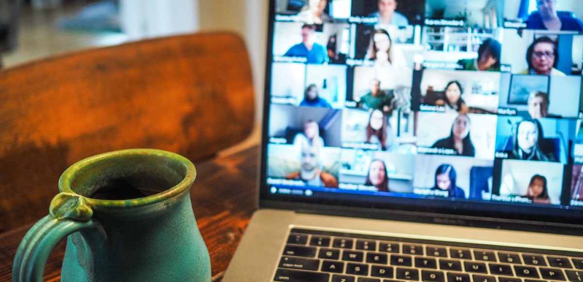 A coffee mug in front of a laptop with a zoom chat grid of participants on the screen.