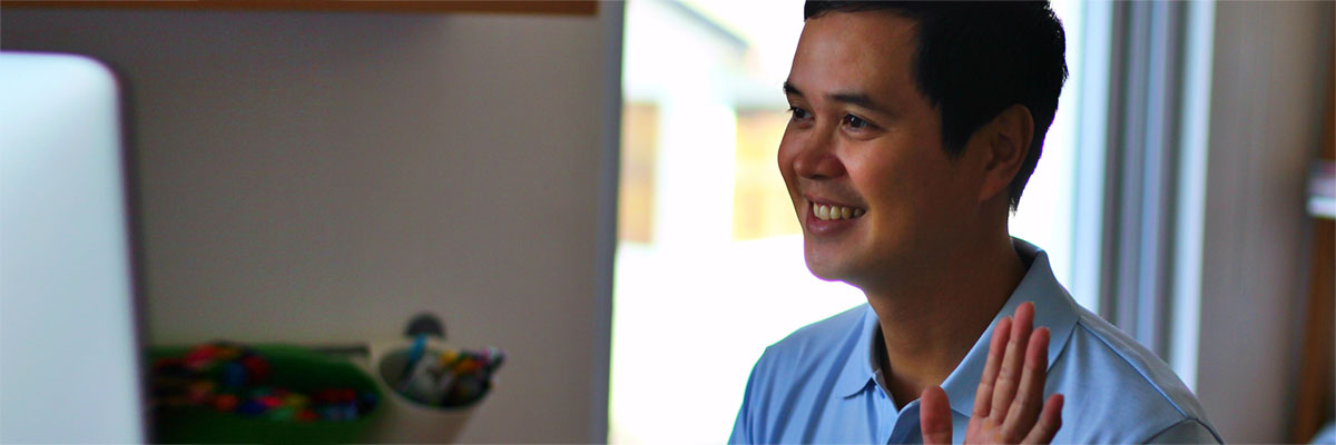 A man in front of his computer smiling and waving.
