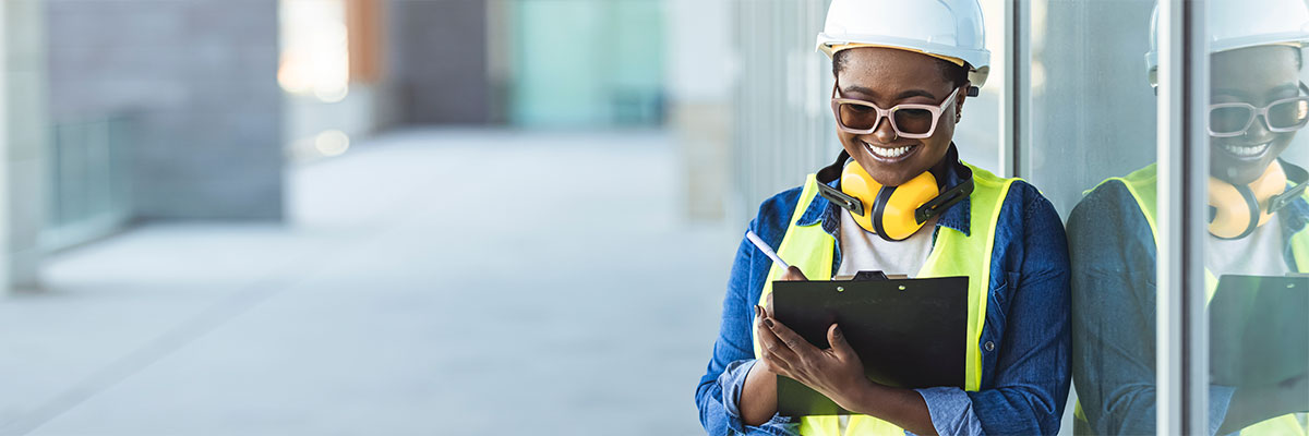 A smiling Black woman in PPE is carrying a clipboard and making notes for an inspection report.