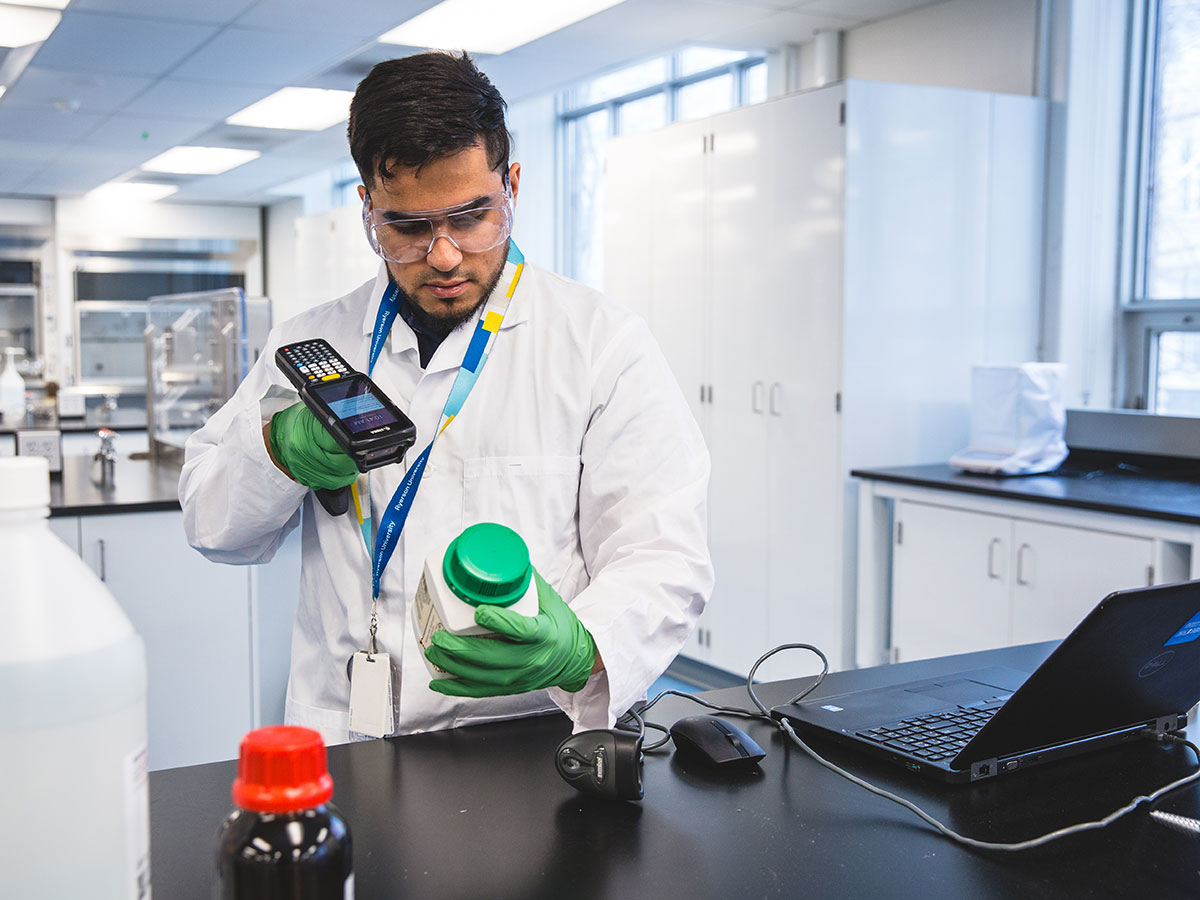 A lab technician with gloves and safety glasses scans a storage container of chemicals.