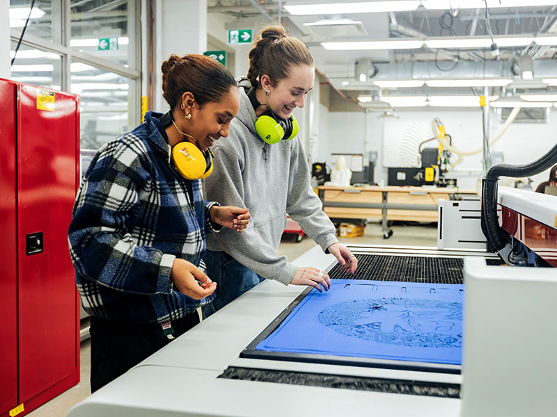 Students with hearing protection work in the Design + Technology Lab with a plotter that cut shapes from a substrate