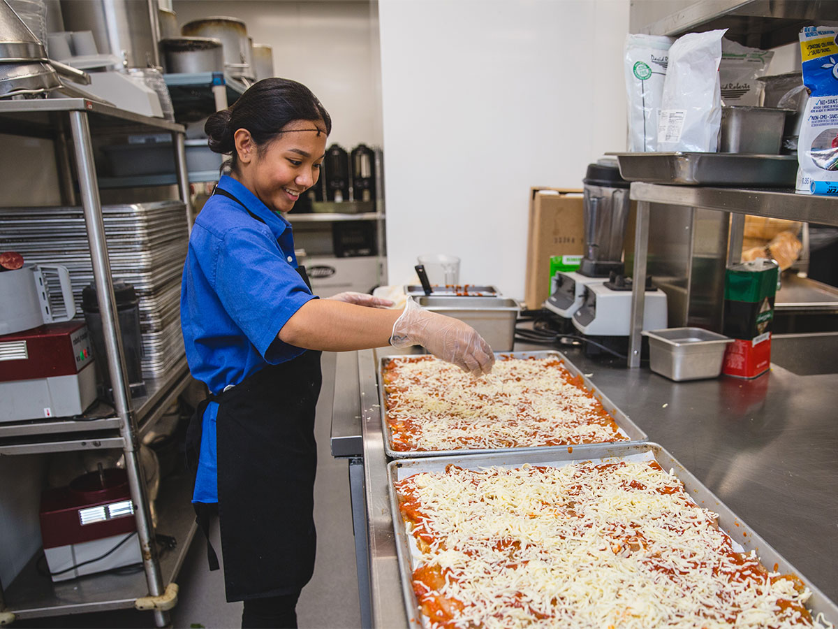 A female Food Services worker wearing an apron, gloves and a hair net sprinkles shredded cheese over food to be baked.