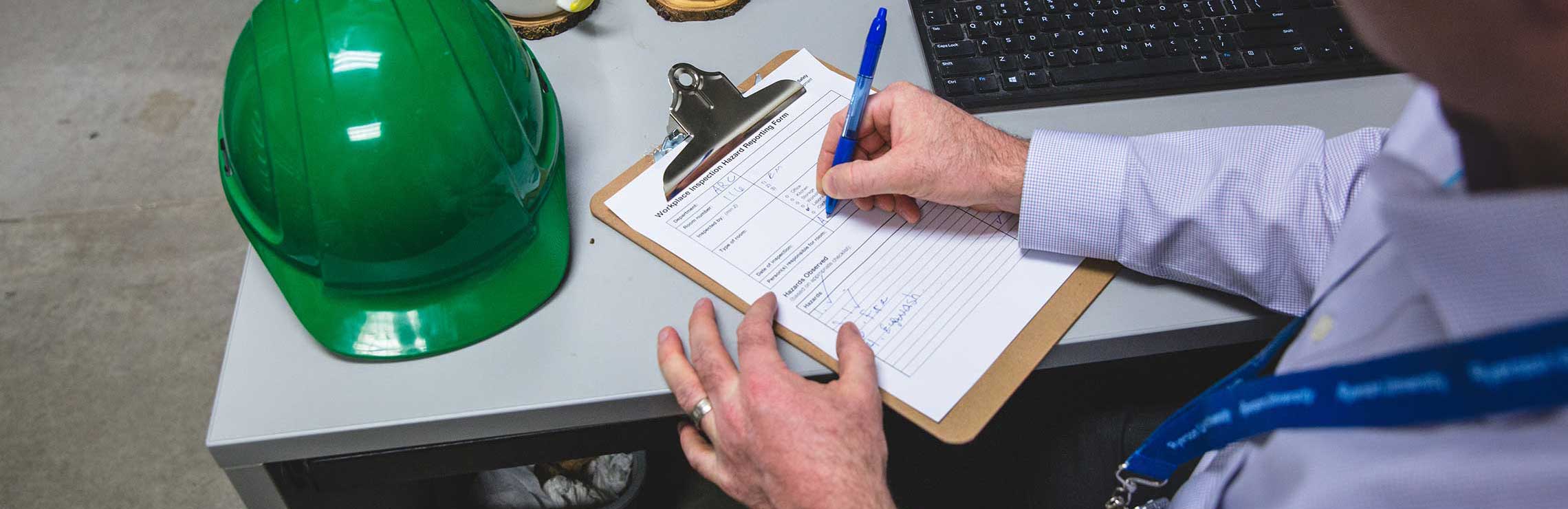 An inspector's hands fill out an inspection form at his desk with his hard hat next to the clipboard.