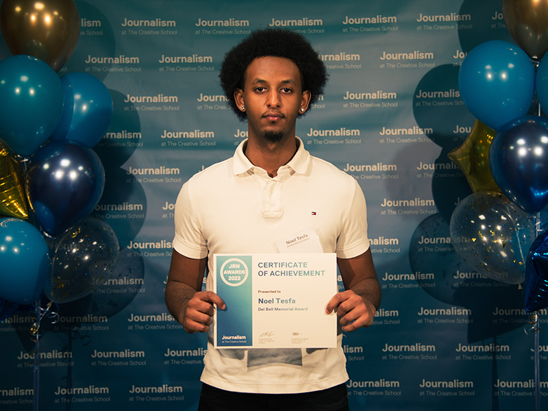 Noel Tesfa holds a certificate while standing in front of a blue backdrop that repeats, "Journalism at The Creative School."