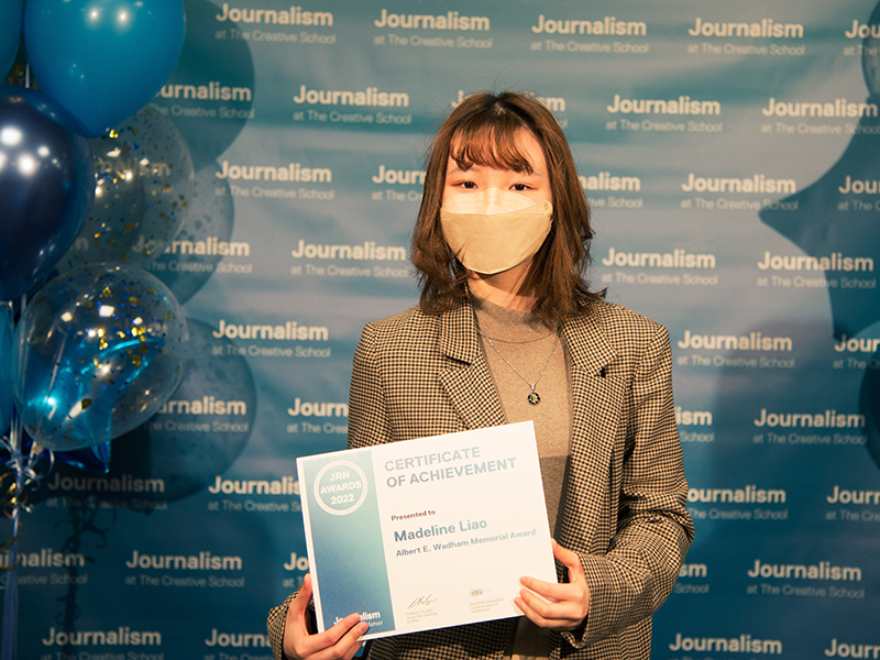 Madelina Liao holds a certificate while standing in front of a blue backdrop that repeats, "Journalism at The Creative School."