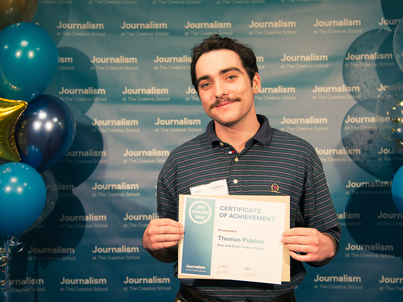 Thomas Publow holds a certificate while standing in front of a blue backdrop that repeats, "Journalism at The Creative School."