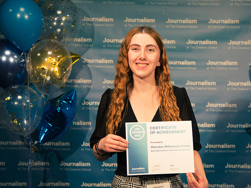 Sheridan Williamson-Fraser holds a certificate while standing in front of a blue backdrop that repeats, "Journalism at The Creative School."