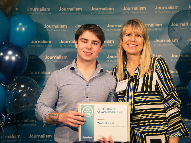 Maxwell Loslo holds a certificate while standing with Diana Spremo in front of a blue backdrop that repeats, "Journalism at The Creative School."