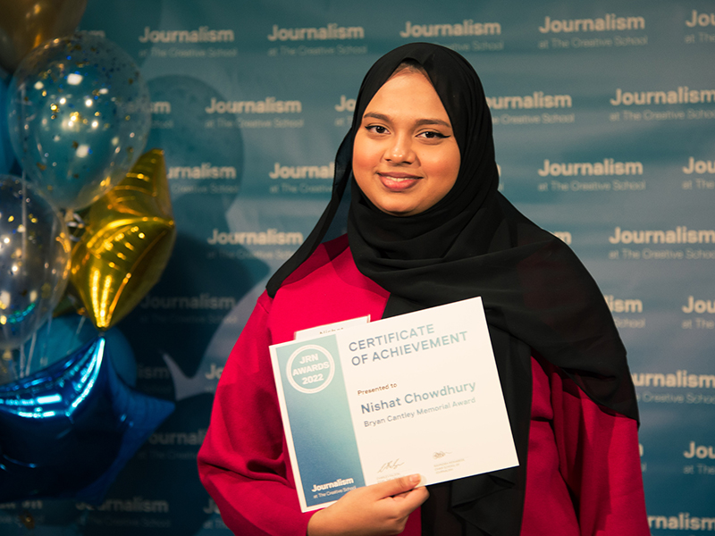 Nishat Chowdhury holds a certificate while standing in front of a blue backdrop that repeats, "Journalism at The Creative School."