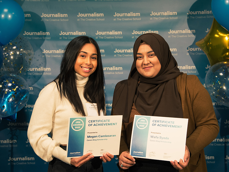 Megan Camlasaran and Wafa Syeda hold certificates while standing in front of a blue backdrop that repeats, "Journalism at The Creative School."