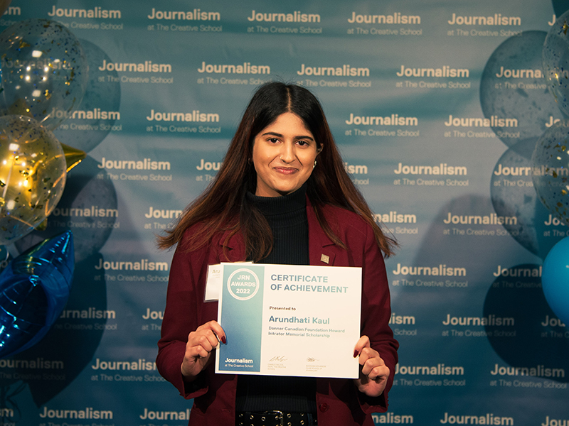Arundhati Kaul holds a certificate while standing in front of a blue backdrop that repeats, "Journalism at The Creative School."