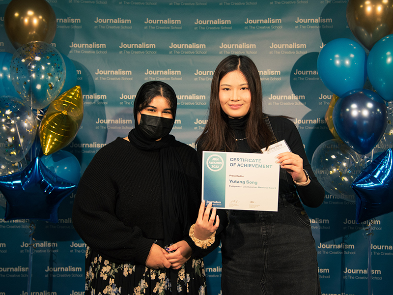 Yutang Song holds a certificate while standing with Abeer Khan in front of a blue backdrop that repeats, "Journalism at The Creative School."