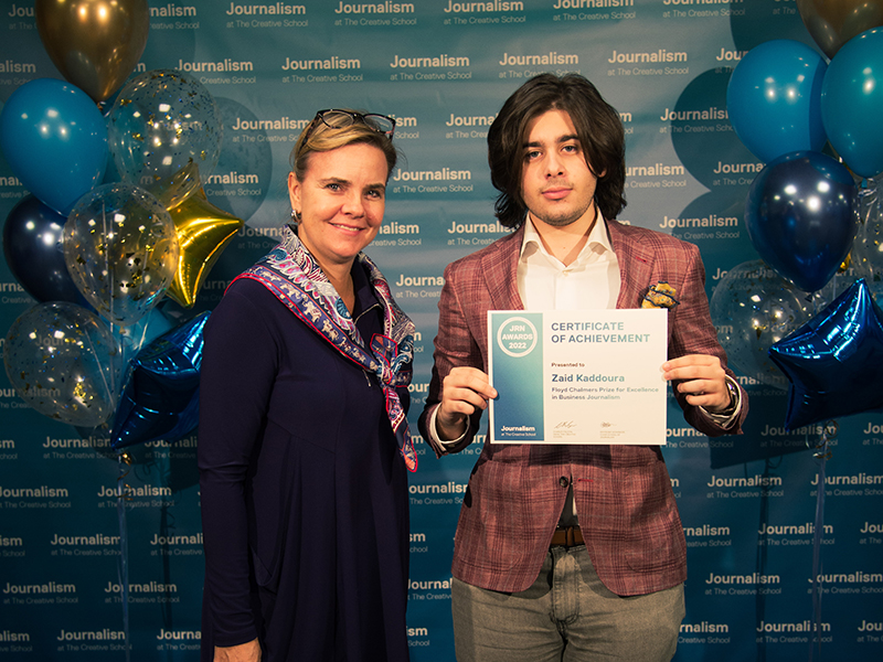 Zaid Kaddoura a holds a certificate while standing with Michelle Robitaille in front of a blue backdrop that repeats, "Journalism at The Creative School."