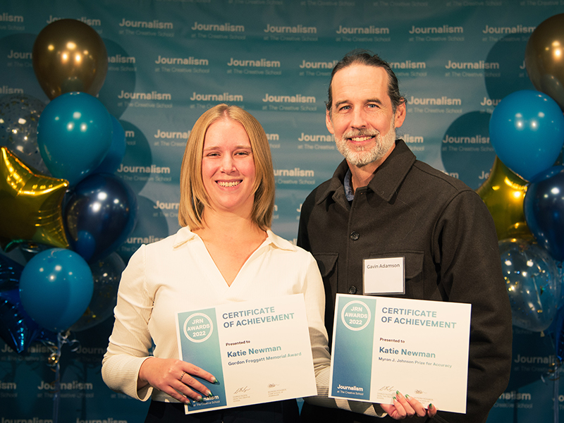 Katie Newman holds a certificate with Gavin Adamson while standing in front of a blue backdrop that repeats, "Journalism at The Creative School."