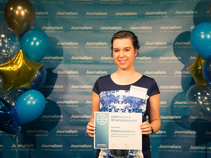 Charlene Hatcher holds a certificate while standing in front of a blue backdrop that repeats, "Journalism at The Creative School."
