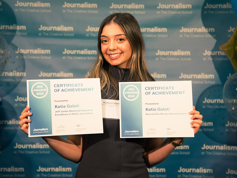 Katia galati holds two certificates while standing in front of a blue backdrop that repeats, "Journalism at The Creative School."