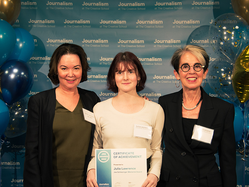 Julia Lawrence holds a certificate while standing with Joanne McPherson and Judith Levene in front of a blue backdrop that repeats, "Journalism at The Creative School."