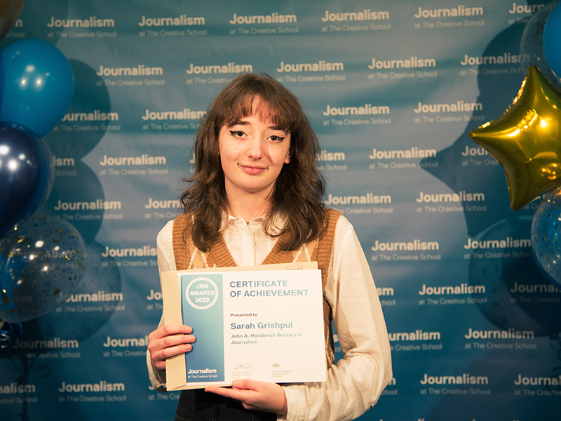 Sarah Grishpul holds a certificate while standing in front of a blue backdrop that repeats, "Journalism at The Creative School."
