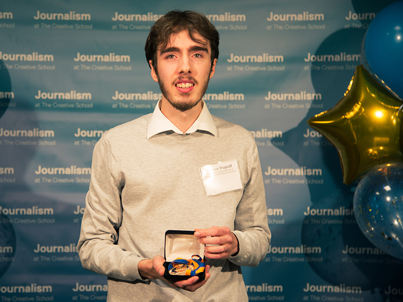 Trevor Popoff holds a gold medal while standing in front of a blue backdrop that repeats, "Journalism at The Creative School."