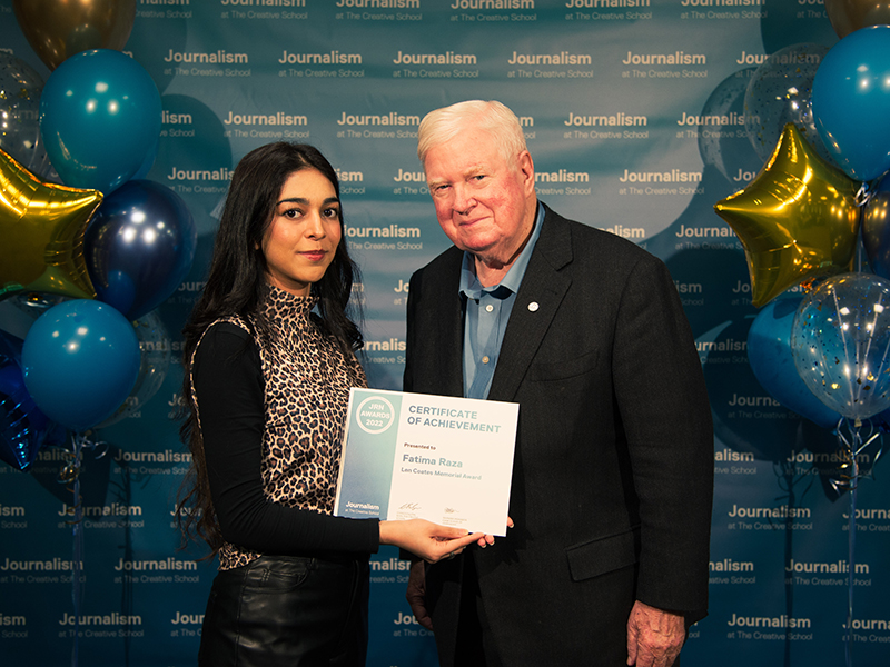 Fatima Raza holds a certificate while standing with Hugh Scully in front of a blue backdrop that repeats, "Journalism at The Creative School."