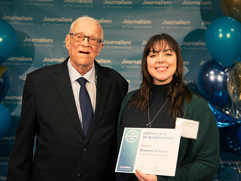 Breanna Schnurr holds a certificate while standing with Norris MacDonald in front of a blue backdrop that repeats, "Journalism at The Creative School."