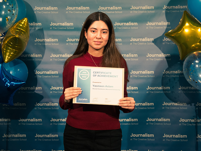 Yasmeen Aslam holds a while standing in front of a blue backdrop that repeats, "Journalism at The Creative School."